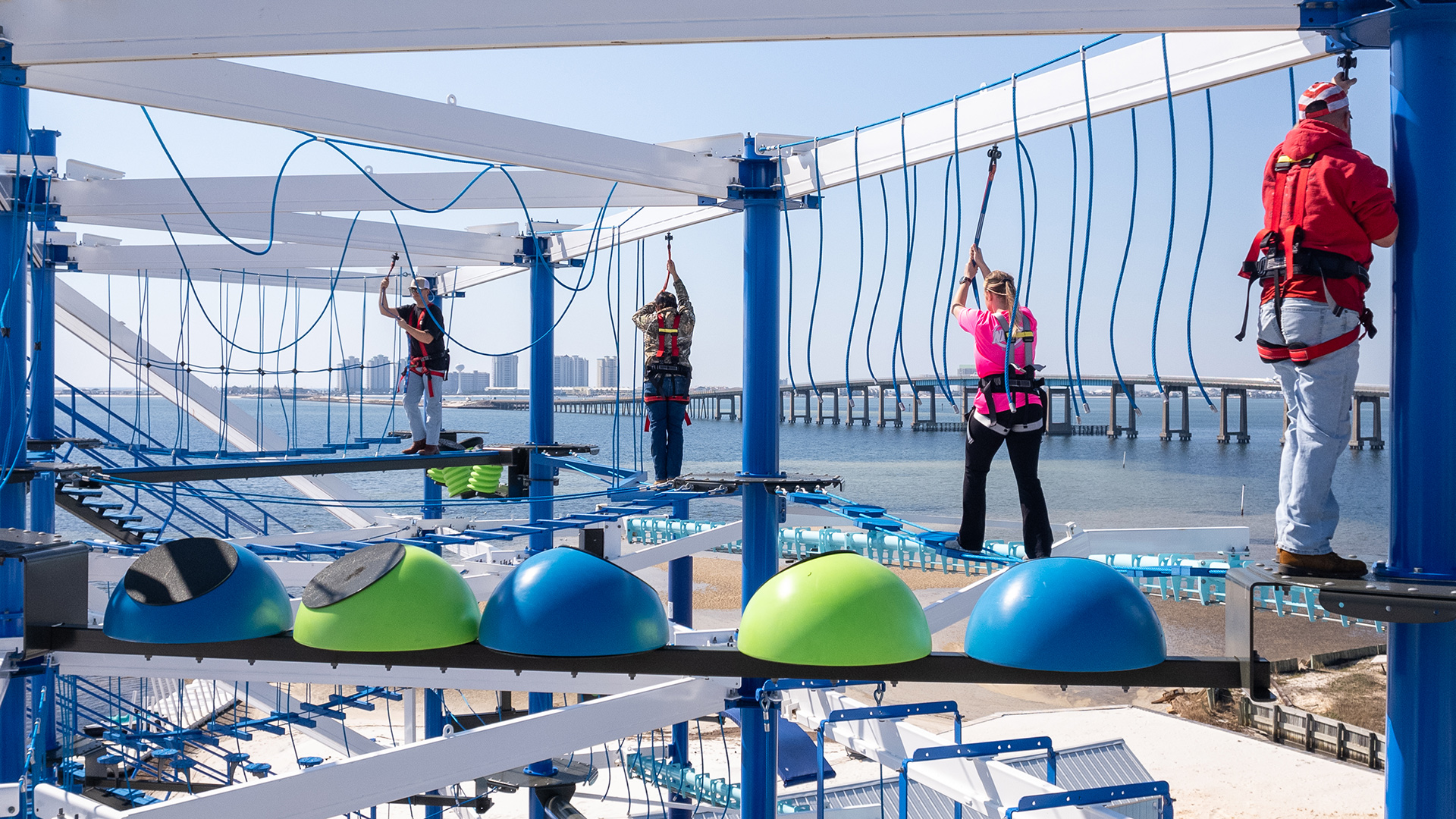 Four people balance on different obstacles on the top level of Navarre Family Watersports' ropes course with Navarre Bridge and beach in the background